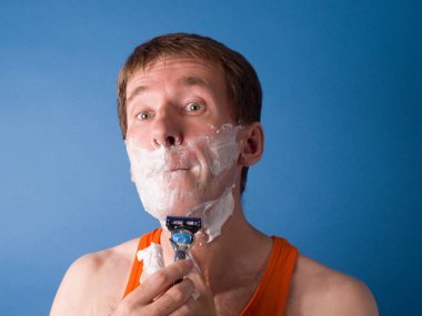 Yakın plan. Turuncu gömlekli adam dikkatlice tıraş oluyor. Mavi arkaplanda stüdyo fotoğrafçılığı