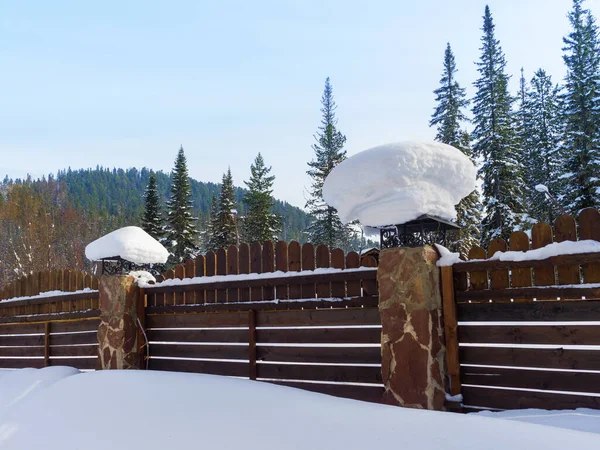 Stylish wooden fence with stone pillars. Large clumps of snow lie on the forged roofs of the pillars