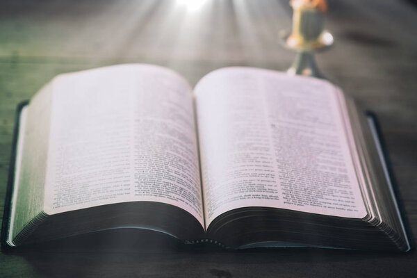 Opened Holy Bible book on wooden table against window light.