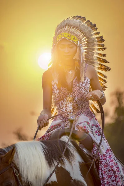 Bohem Kız gün batımında ahırda atını sürüyor. Güzel kırmızı kahverengi at ve kızılderili kadınlar savaş şapkalı güzellik portresi yerli Amerikan tüylü başlık.