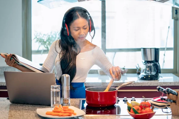 Mutlu kadın operatör danışmanlık hizmeti, video konferansında kulaklık ve mikrofonla konuşarak meşgul. Kırmızı tencerede yemek pişirirken mutfakta bir dizüstü bilgisayar. Ev kavramından çalış.