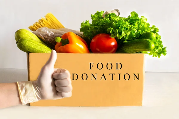 Food delivery. Food donation on table. Help cardboard box wiht various canned food, cereals, pasta and vegetables. Volunteer box. Female hand in hygienic gloves