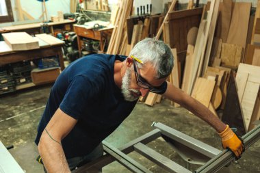 Gözlüklü marangoz ve gri saçlı kesme makinesini ahşap atölyesinde ayarlıyor..