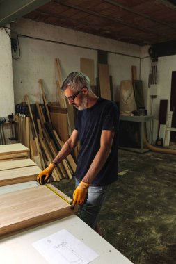 Marangoz bir tahta parçasını ahşap atölyesinde mezura ile ölçüyor.