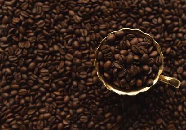 Goldene Tasse auf Untertasse gefüllt mit Kaffeebohnen auf verschwommenem Hintergrund voller Kaffeebohnen — Stockfoto