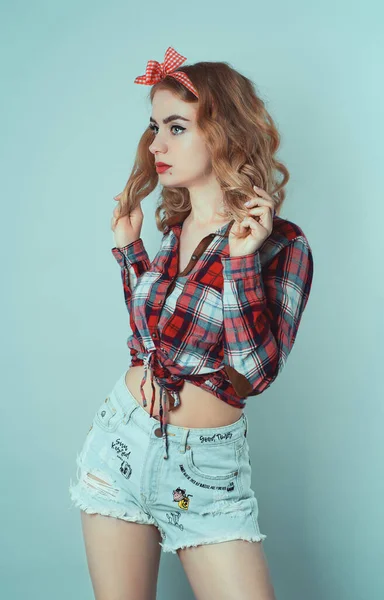 Pin-up Mädchen mit lockigem blondem Haar mit rot karierter Schleife und Hemd in Jeans-Shorts. — Stockfoto