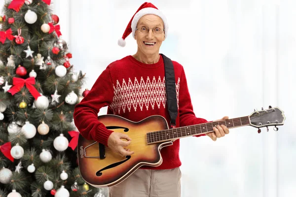 Sênior tocando uma guitarra acústica na frente de uma árvore de natal — Fotografia de Stock