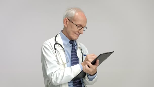 Ouderen dokter schrijven op een Klembord en glimlachen — Stockvideo