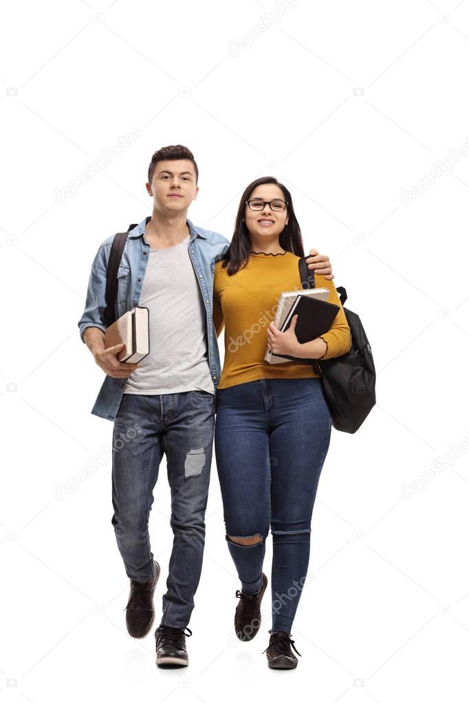 Full length portrait of two teenage students with backpacks and books walking towards the camera isolated on white background