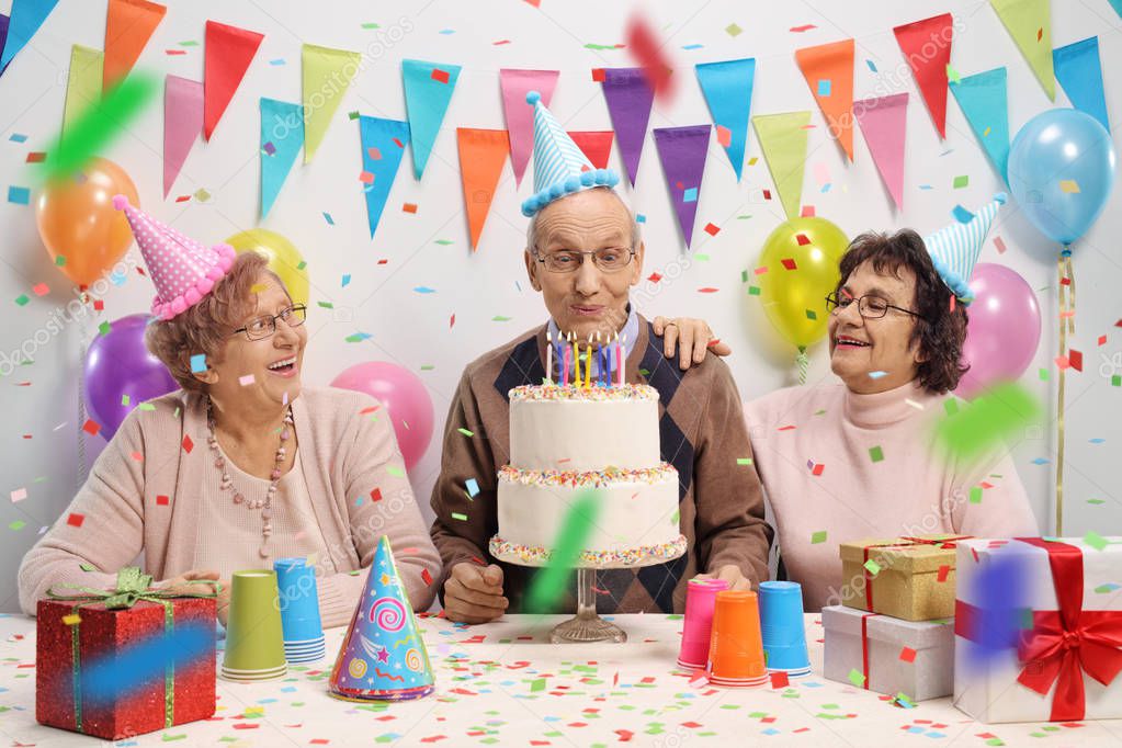 Elderly man with two elderly women blowing candles on a birthday cake against a wall with decoration flags and balloons