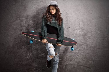 Paslı bir gri duvara yaslanmış bir longboard ile genç kız