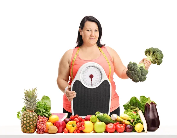 超重妇女持有体重秤和西兰花哑铃在桌子后面的水果和蔬菜隔离在白色背景 — 图库照片