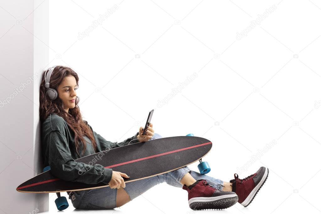 Teenage skater girl with a longboard leaning against a wall and listening to music on a phone isolated on white background