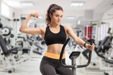 Fitness woman riding an exercise bike and flexing her biceps in a gym