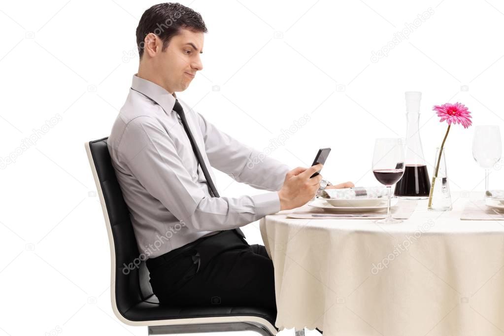 Disappointed young man seated at a restaurant table looking at his phone isolated on white background