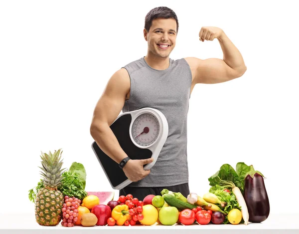 年轻人在运动装重量秤和弯曲他的二头肌在桌后面与水果和蔬菜隔绝在白色背景下 — 图库照片