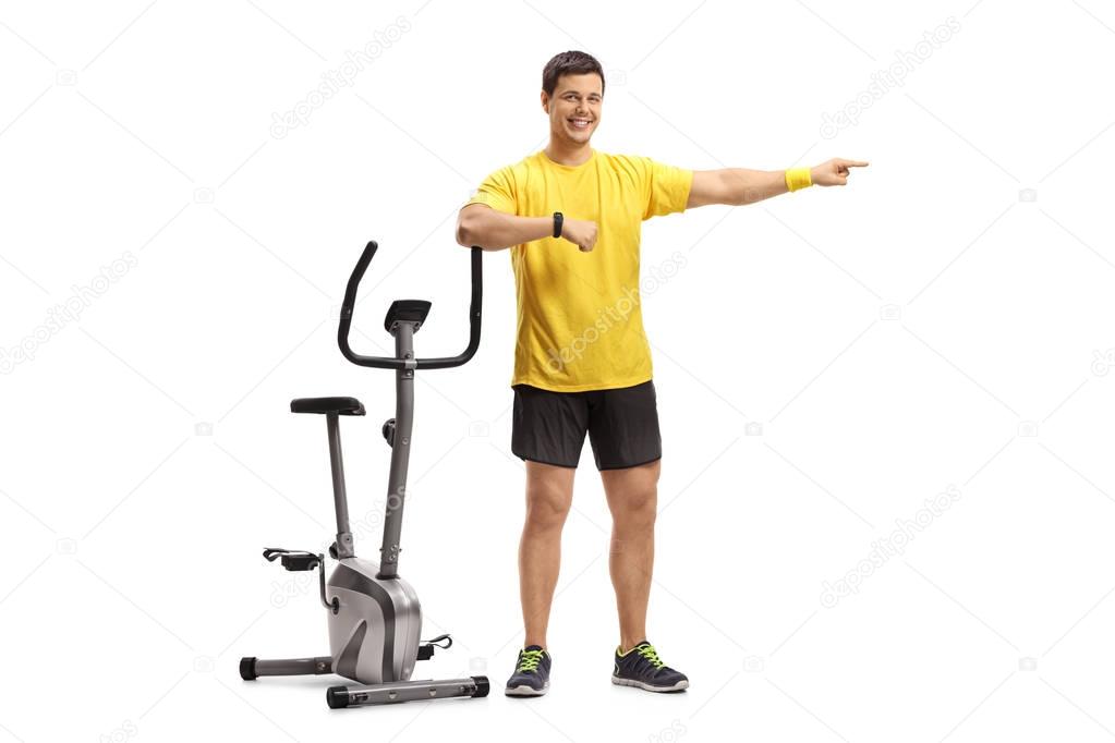 Full length portrait of a young man leaning on an exercise bike and pointing isolated on white background