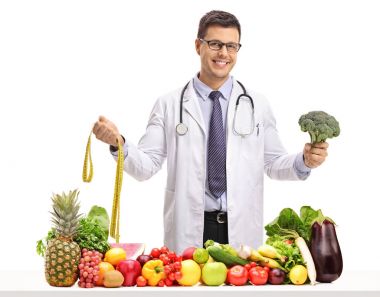 Bir ölçüm bandı ve brokoli ile meyve ve sebze beyaz arka plan üzerinde izole bir masanın arkasında tutarak doktor
