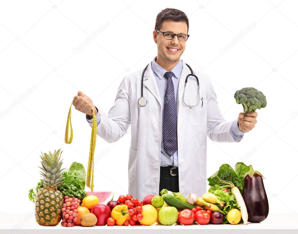 Doctor holding a measuring tape and broccoli behind a table with fruit and vegetables isolated on white background