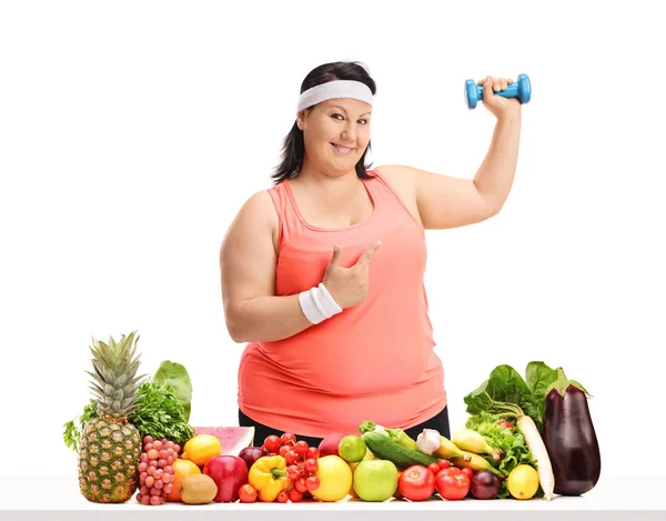 太りすぎの女性の小さなダンベルを押しながら背後の果物や野菜は 白い背景で隔離のテーブルを指す — ストック写真