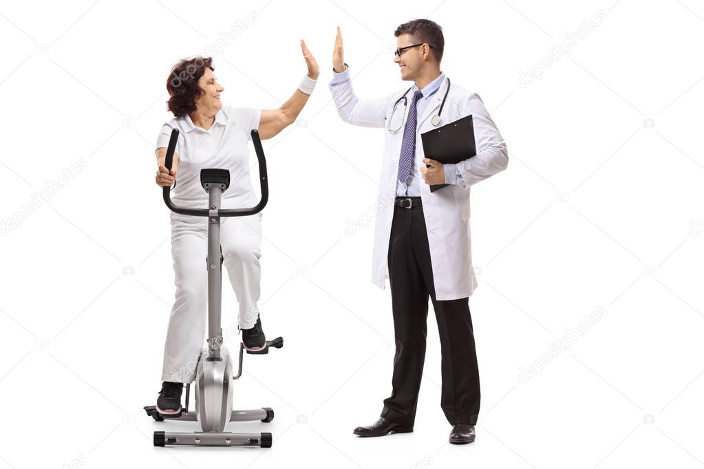 Elderly woman riding an exercise bike and high-fiving a doctor isolated on white background