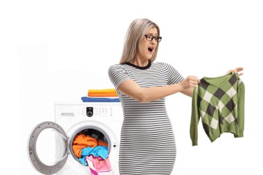 Shocked young woman holding a shrunken blouse in front of washing machine isolated on white background clipart