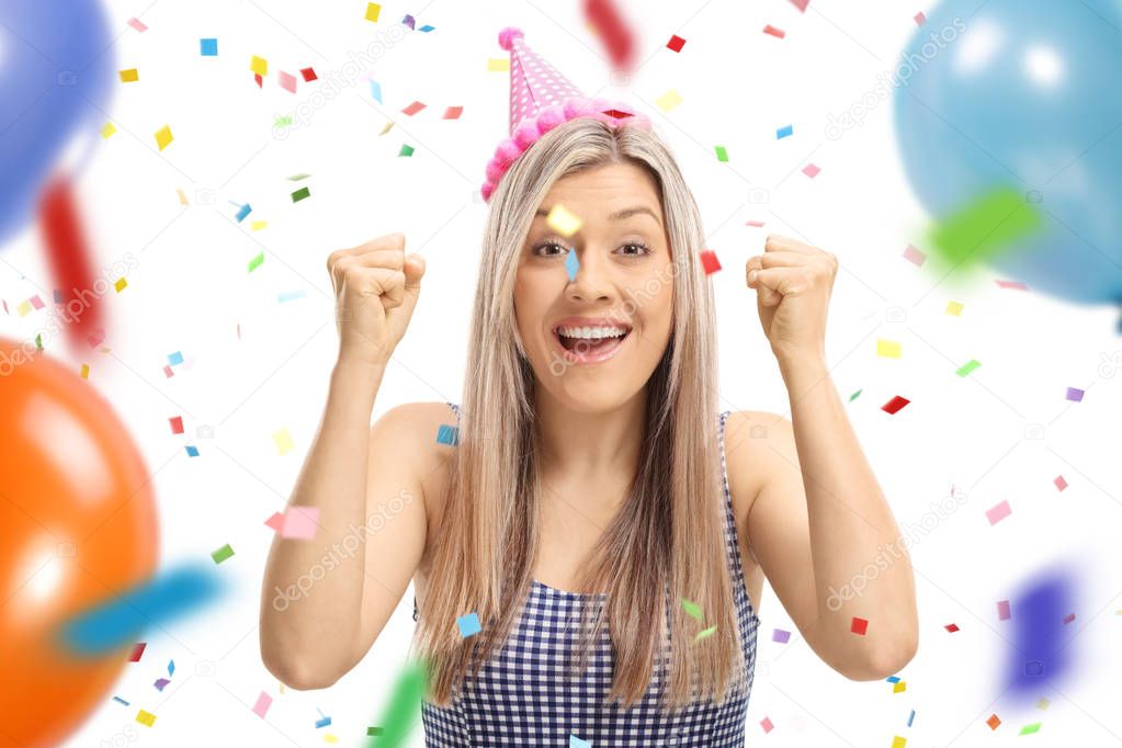 Young woman wearing a party hat gesturing happiness with confetti streamers and balloons flying around her isolated on white background