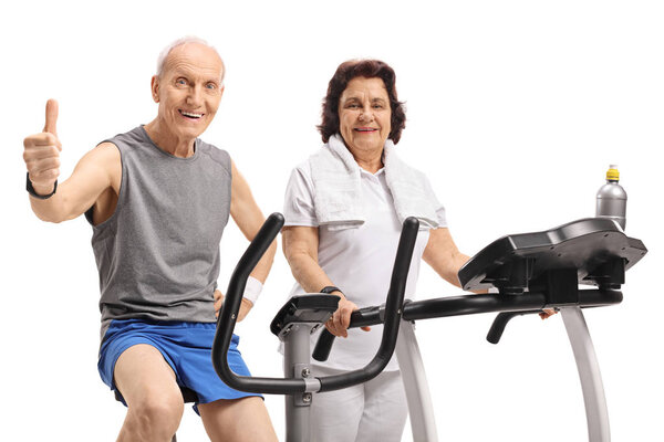 Elderly man on an exercise bike making a thumb up sign with an elderly woman on a treadmill isolated on white background
