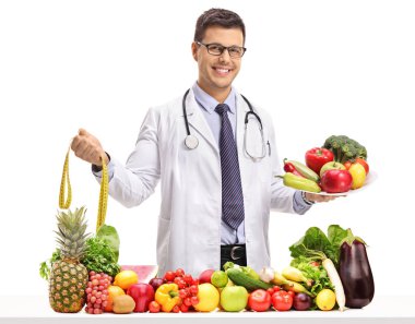 Bir ölçüm bandı ve bir tabak meyve ve sebze beyaz arka plan üzerinde izole bir masanın arkasında tutarak doktor