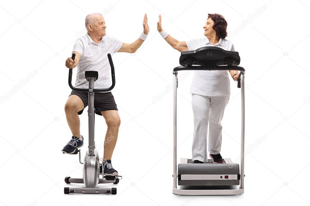 Elderly man exercising on a stationary bike and an elderly woman on a treadmill high-fiving each other isolated on white background