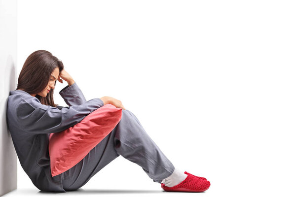 Грустная молодая женщина в пижаме сидит на полу и держит подушку изолированы на белом фоне
