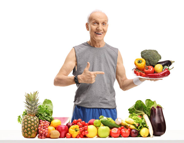 Senior behind a table with fruit and vegetables holding a plate and pointing isolated on white background