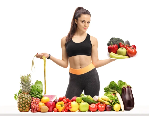 健身女孩拿着一个盘子和一个测量带在桌子后面的水果和蔬菜隔离在白色背景 — 图库照片