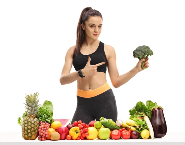 健身妇女在桌子后面的水果和蔬菜持有花椰菜和指向孤立的白色背景 — 图库照片