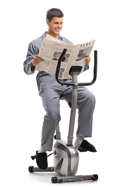 白い背景に分離されたエアロバイクで新聞を読んでパジャマ姿で若い男 — ストック写真