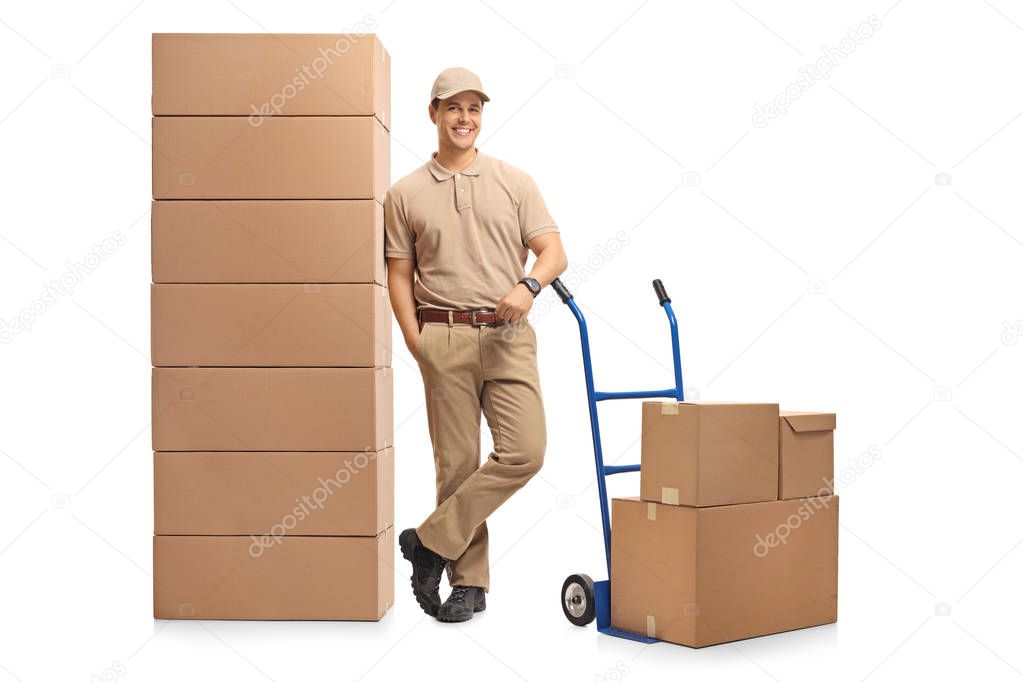 Full length portrait of a delivery man with a hand truck leaning on a pile of boxes isolated on white background