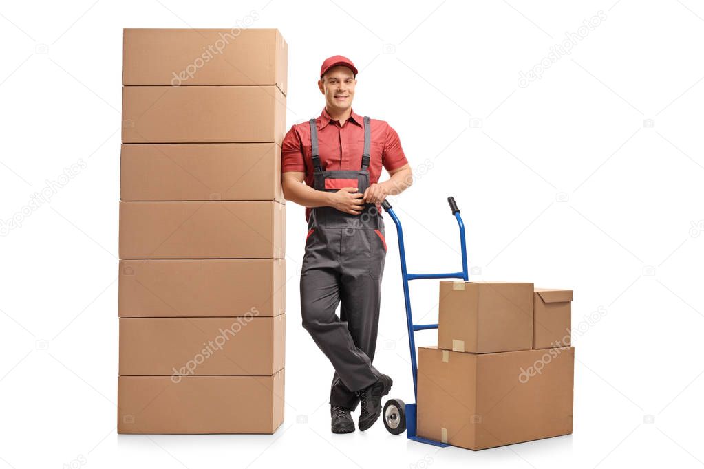 Full length portrait of a mover with a hand truck leaning on a stack of boxes and smiling isolated on white background