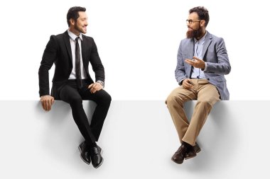 Takım elbiseli bir adam ve sakallı bir adam boş bir panelde oturuyor.