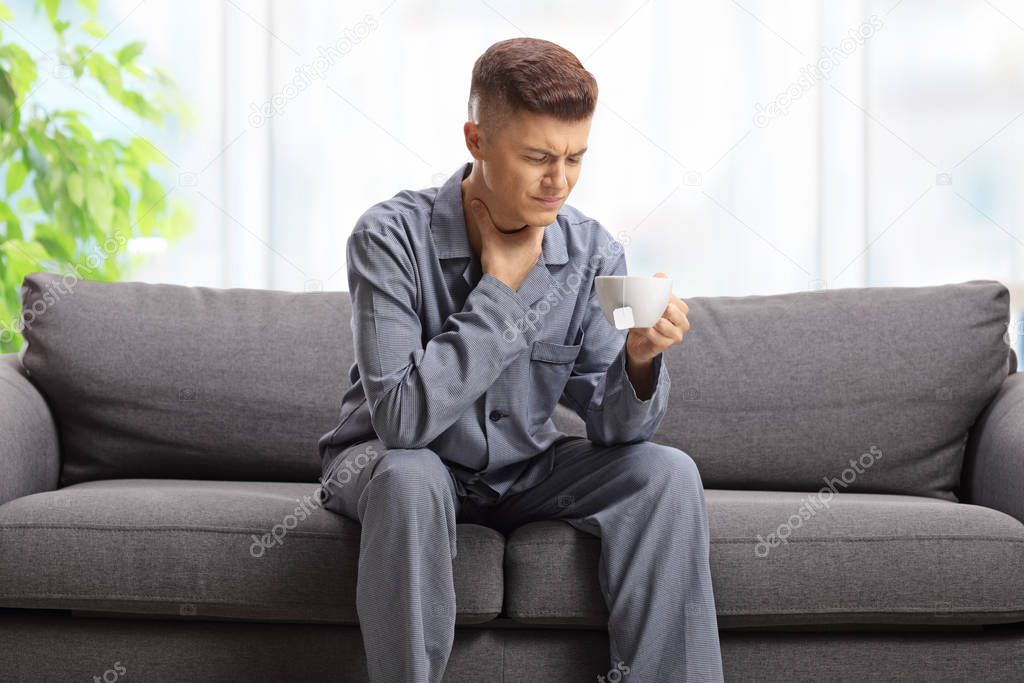 Young man in pyjamas feeling unwell, holding his throat and drin