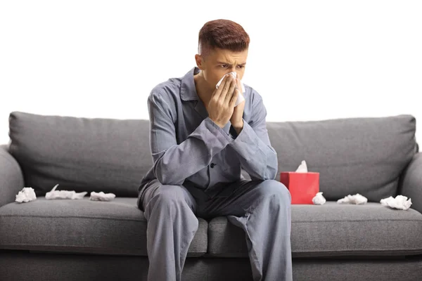 Guy in pyjamas sitting on a couch and blowing nose — Stock Photo, Image
