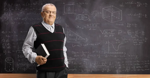 Professor idoso com um livro em pé em frente a um quadro negro — Fotografia de Stock