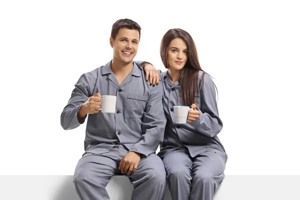 Young couple in pyjamas holding coffee mugs and sitting on a pan — Stock Photo, Image