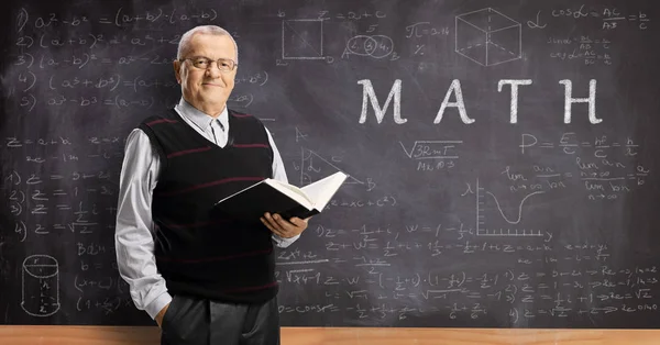 Elderly male teacher holding a book in front of a math blackboar — Stock Photo, Image