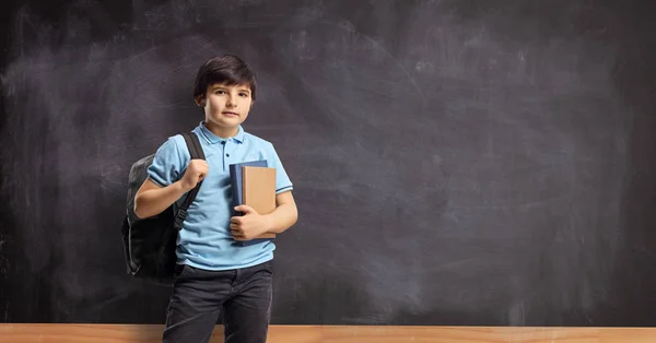 Kid with a backpack and books standing in front of a blackboard — 스톡 사진