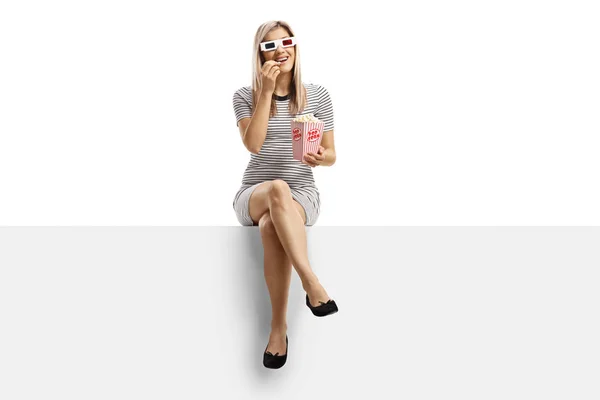 Woman sitting on a blank panel with a box of popcorn and wearing — Stock Photo, Image