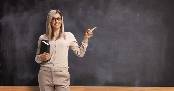 Female teacher with books pointing to a blackboard — Stock Photo, Image