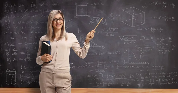 Kobieta nauczyciel trzyma książki i wskazując na tablicę z — Zdjęcie stockowe