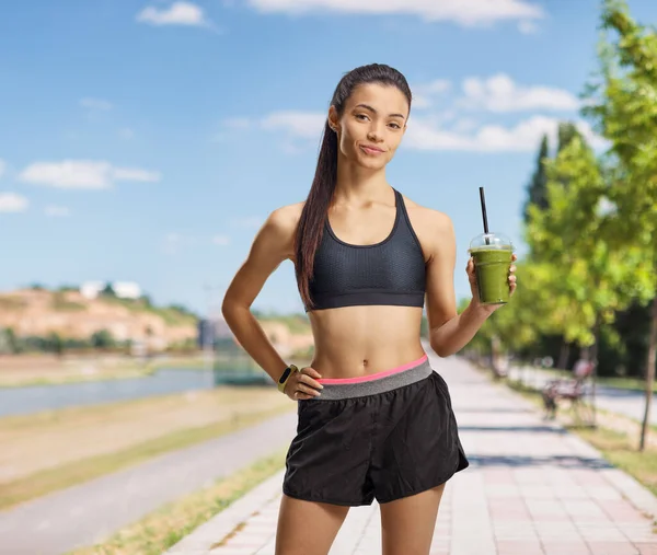 Giovane donna in abbigliamento sportivo che tiene un frullato verde su un jogging — Foto Stock