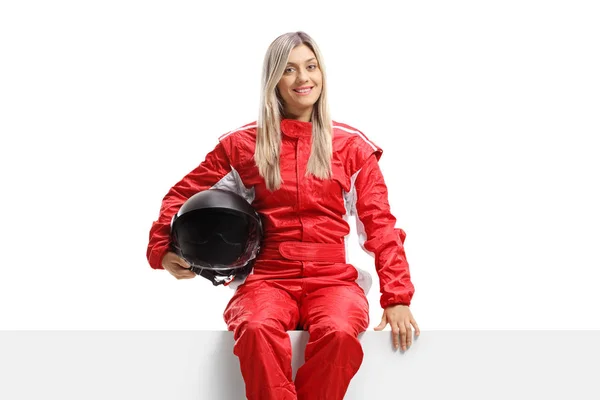 Female racer in a suit holding a helmet and sitting on a panel — Stock Photo, Image