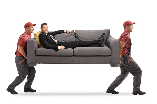 Movers lleva un sofá con un hombre en un traje relajante en el sofá — Foto de Stock
